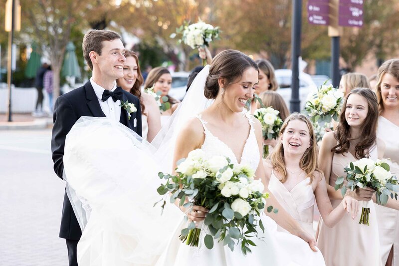 Bride in plunging v neck satin wedding gown with lace detailing holding a romantic whimsical bouquet while she laughs as groom in black tux holds trane of dress with bridal party all around laughing.  Bridesmaids in champagne colored satin dresses.