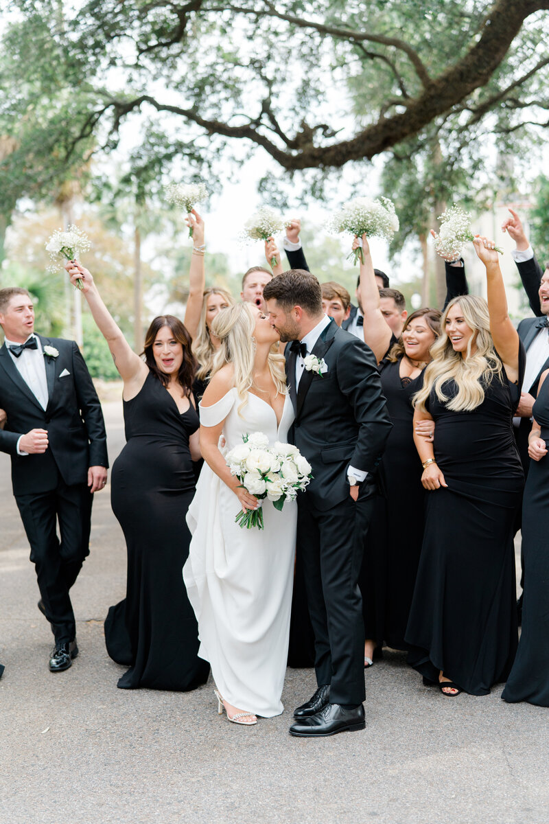 Bridesmaids dressed in black. Bridal party walks on the street with bride and groom with live oak tree in the background
