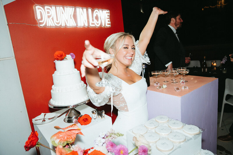 Bride holding up her glass surrounded by the dessert table and a neon sign