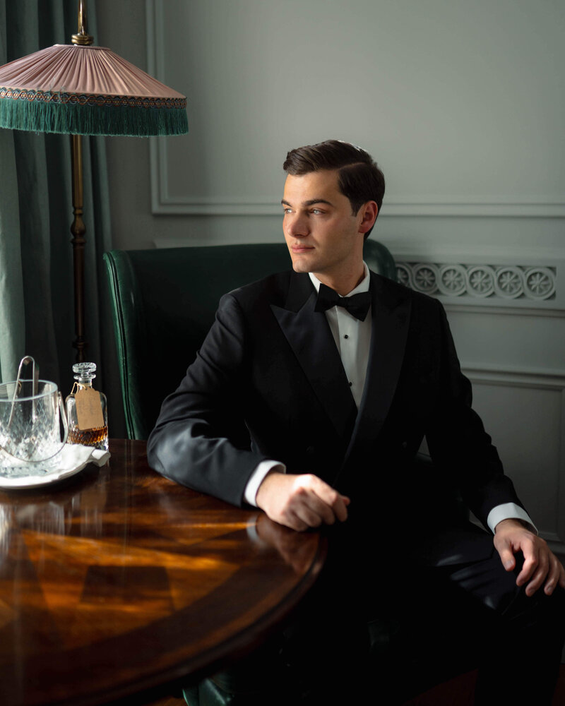 groom wearing a bespoke wedding suit sits at a table looking out the window in his hotel room at the ned hotel before his wedding