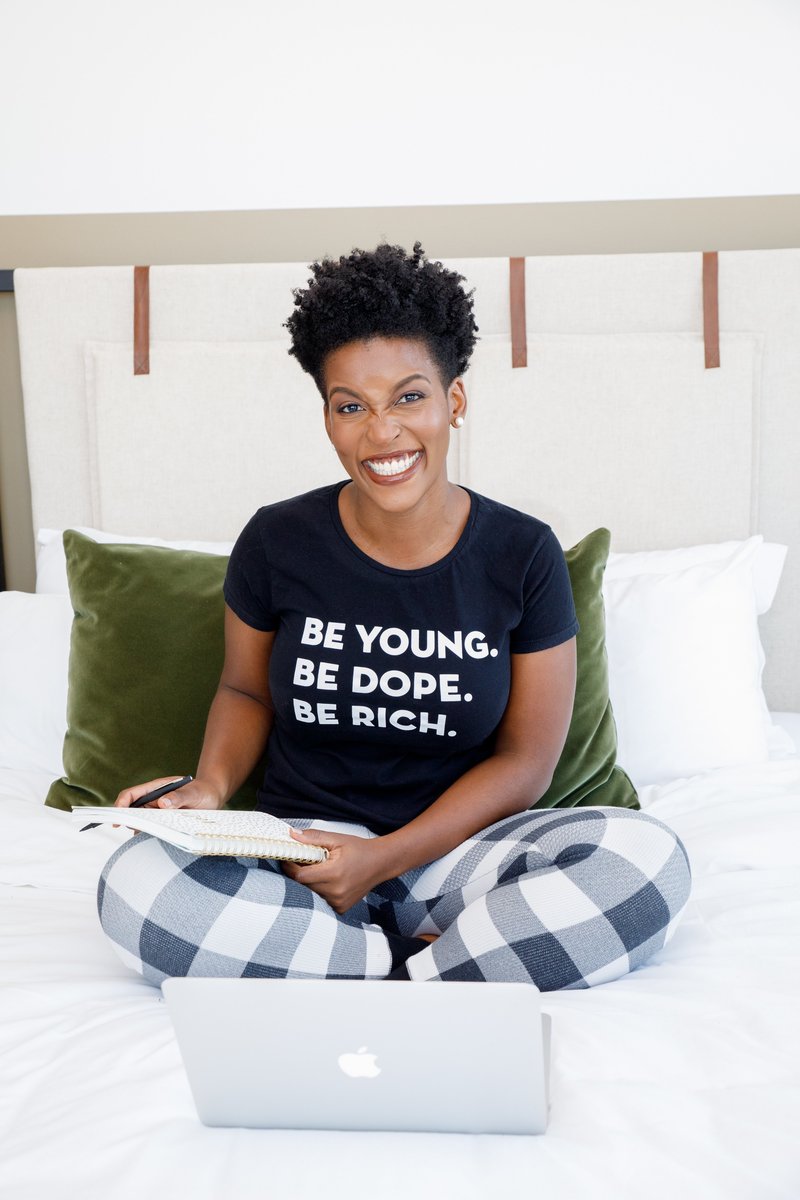 Woman sitting on the bed with a laptop and a Black shirt with the words Be Young Be Rich Be Dope