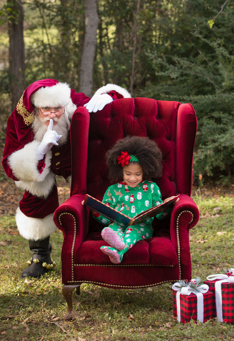 girl-in-holiday-pajamas-sitting-in-santas-red-chair-reading-with-santa-sneaking-in-dalworthing-gardens