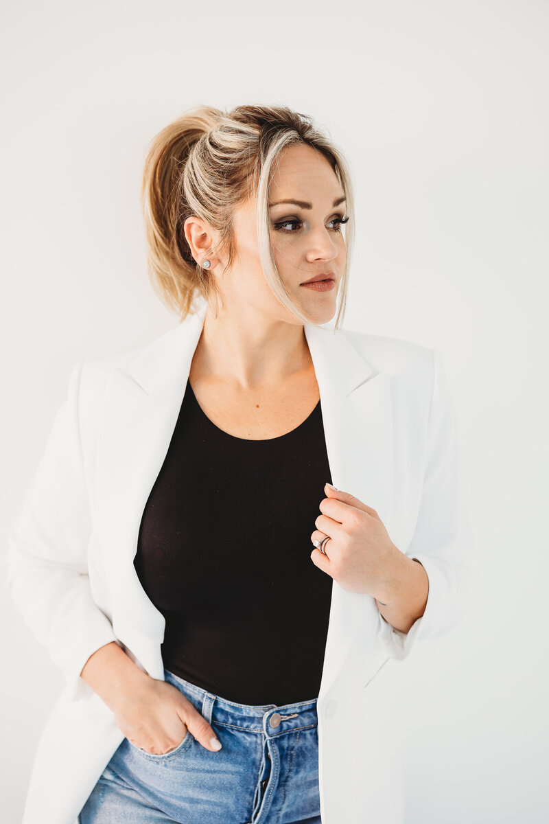 Business woman in white blazer and jeans leaning against a white wall