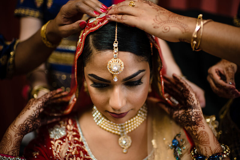 An Indian bride getting her elaborate head jewelry adjusted, with intricate henna designs on her hands and arms, showcasing traditional wedding attire and jewelry.
