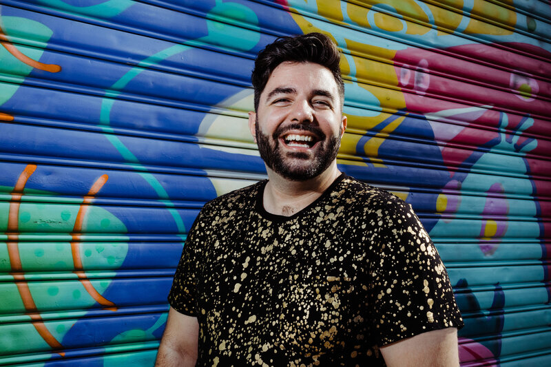 A person smiling while standing in front of a colorful mural.