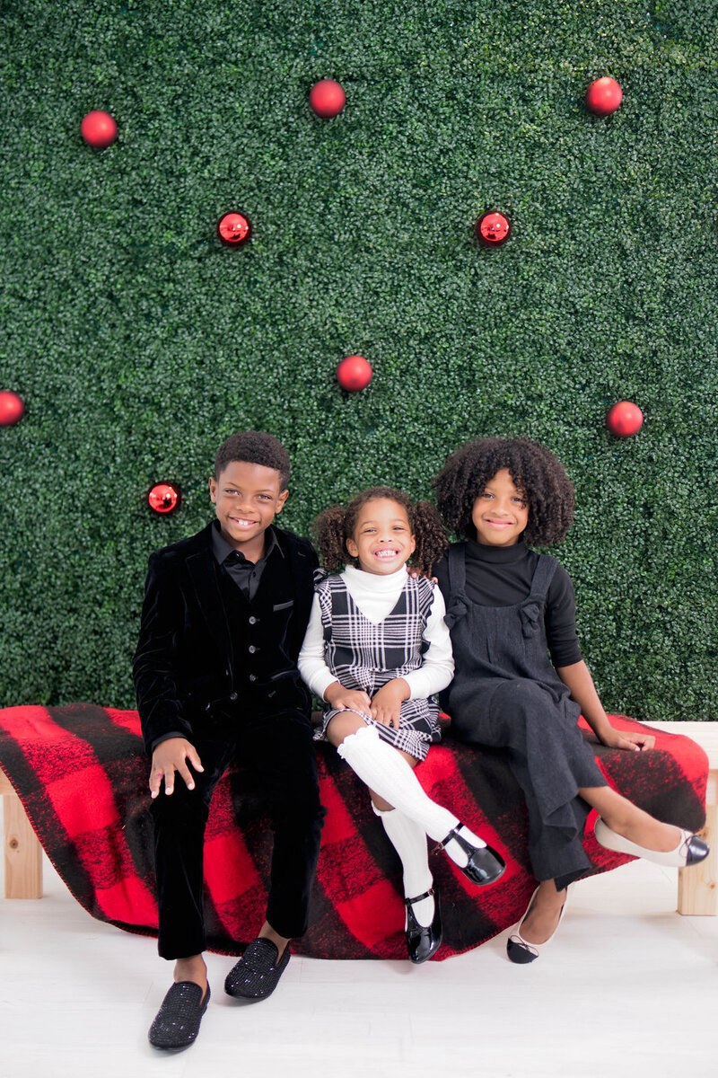 Three children smile at the camera for a Christmas family photoshoot. Behind them is a fir-tree textured wall decorated with red ornaments.