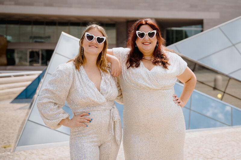 Two brides in heart shaped sun glasses standing together with their arms around each other