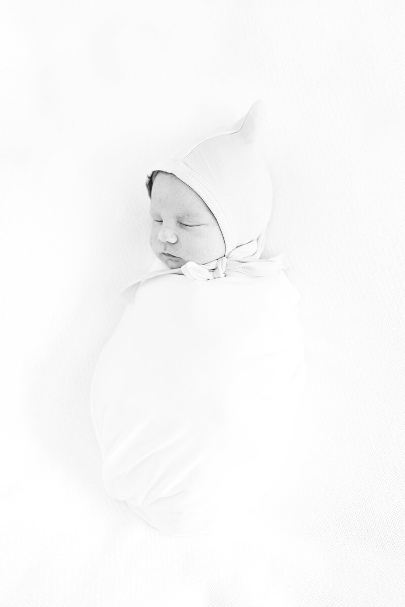 black and white picture of baby boy swaddled with bonnet on and eyes closed during newborn photography session