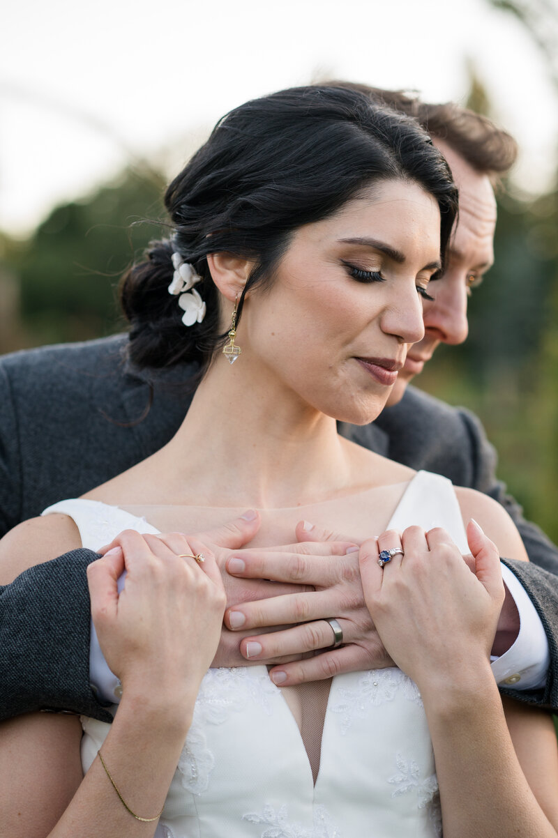 Close-up of a bride being embraced from behind by the groom, both looking serene, with her detailed earring and his ring in focus at Pond House Café