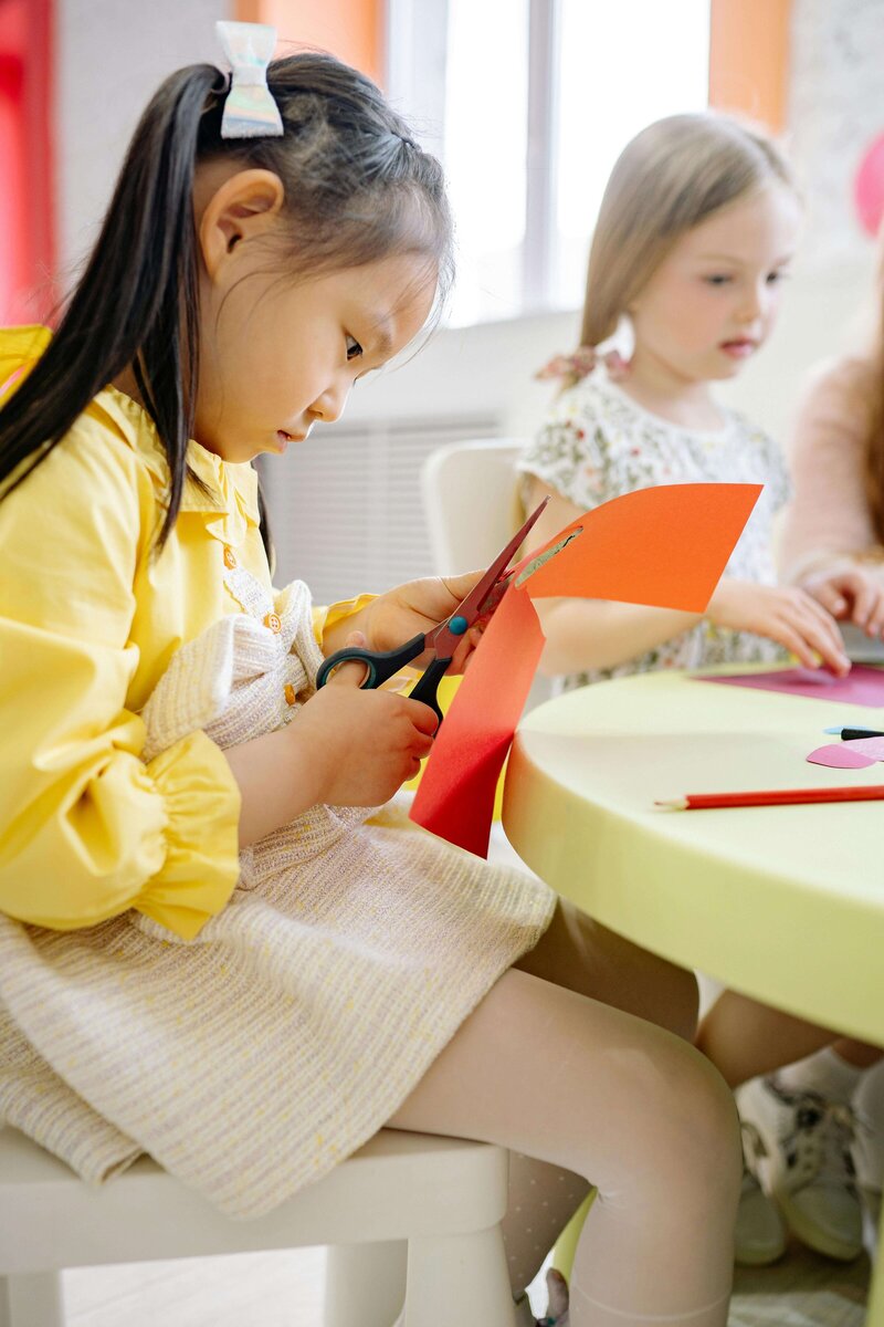 A young girl carefully cuts colorful paper with scissors during an art and craft activity at a table with other children.