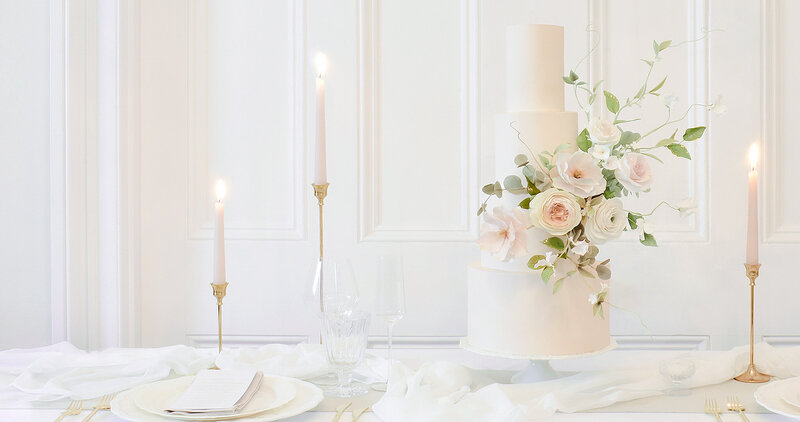 Four tiered wedding cake on a table with tapered candles and styled tableware