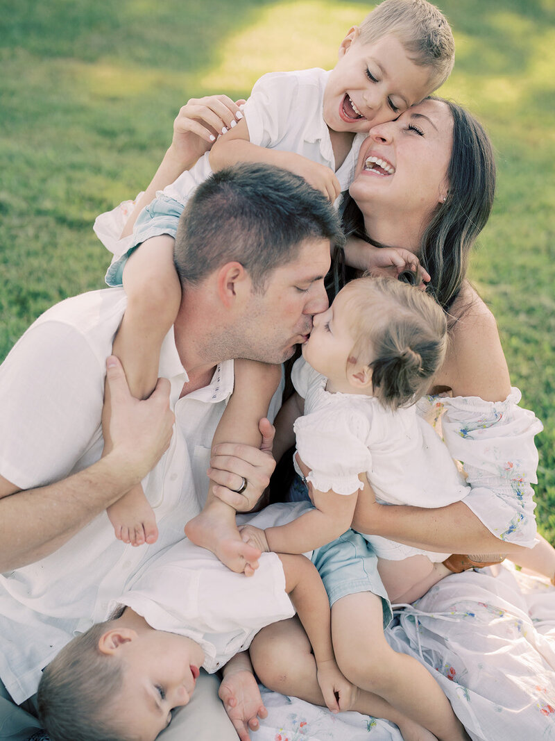Family sits on blanket and lean in to hug and kiss during their Northern Virginia family photo session at Manassas Battlefield.
