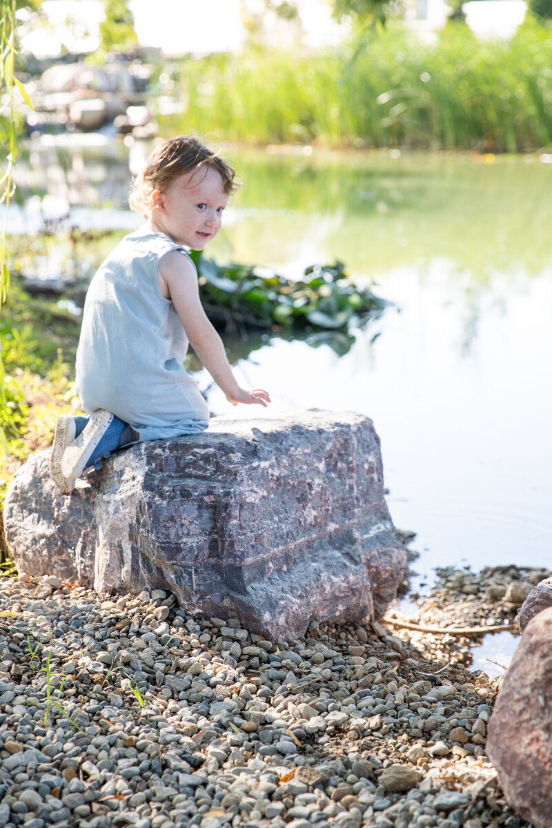 toddler boy kneels on rock next to pond and looks back towards camer