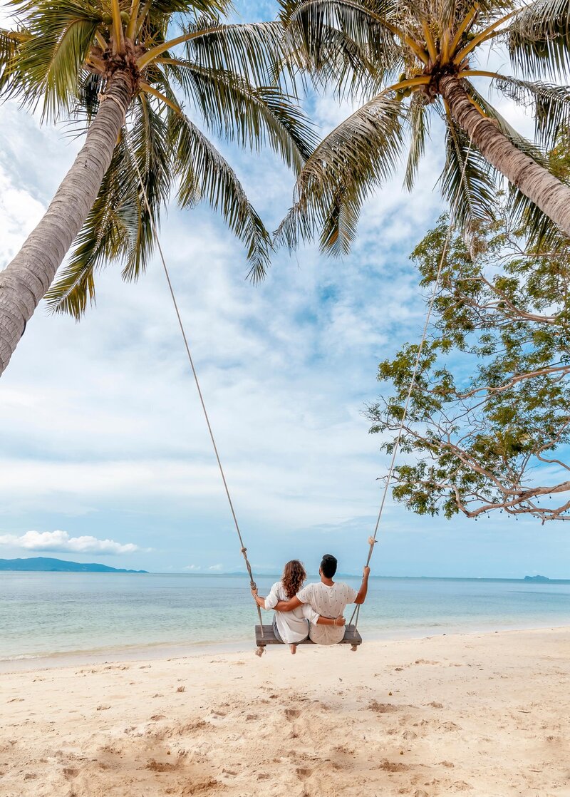A couple sits on a swing hanging from palm trees on a sandy beach, overlooking a calm sea. The sky is partly cloudy, with distant islands visible on the horizon. The scene evokes a peaceful and romantic atmosphere.
