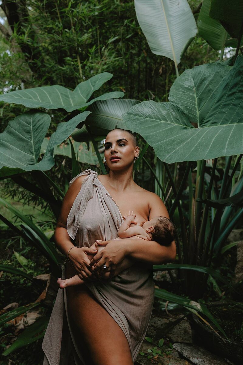 Creative maternity photography with a mother breastfeeding her newborn surrounded by lush tropical greenery.