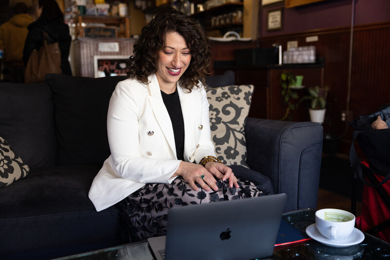 Woman in floral skirt and white blazer sitting on a couch smiling at her open laptop
