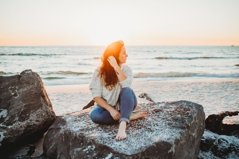 portrait of panama city beach photographer brittney stanley sitting on rock on beach in PCB fl.  She's wearing white sweater and rolled up bluejeans