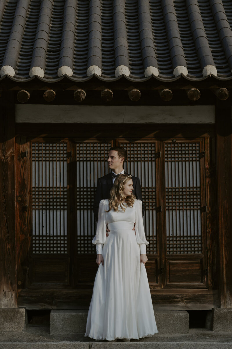 a bride and groom pose in front of a hanok after eloping in seoul