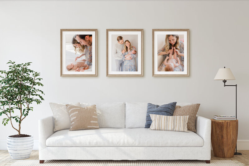 Living room with white couch and neutral pillows, a plant, and a wooden side table. On the wall are three large framed portraits from a newborn session.