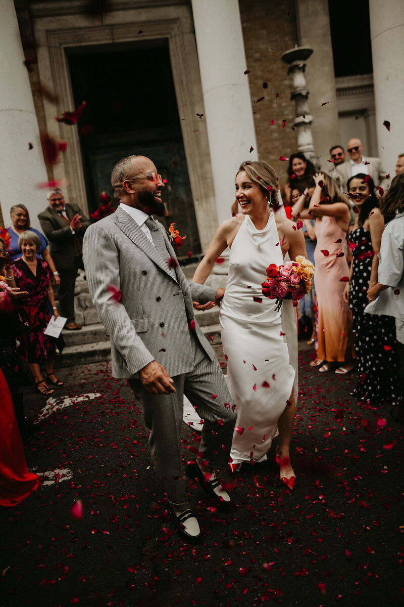 A wedding couple blow pink and red confetti at the camera.