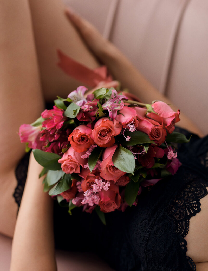 bridal boudoir session of bride to be holding pink bouquet on lap on pink velvet sofa.
