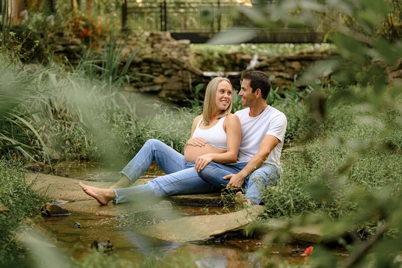 charlotte couple sits in garden dresssed in jeans and white tops during maternity photography session