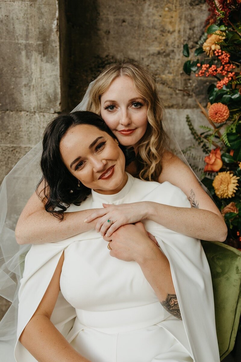 Couple Embracing in Front of Fall Inspired Floral Backdrop and Green Velvet Chair - Megan & Amber | The Ruins PNW Inspired Wedding Hood River Oregon – LGBTQ