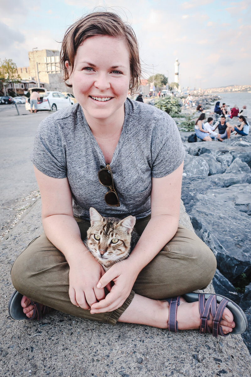 Michelle-Marie Gilkeson smiles  with a cat in her lap by the sea in Turkey