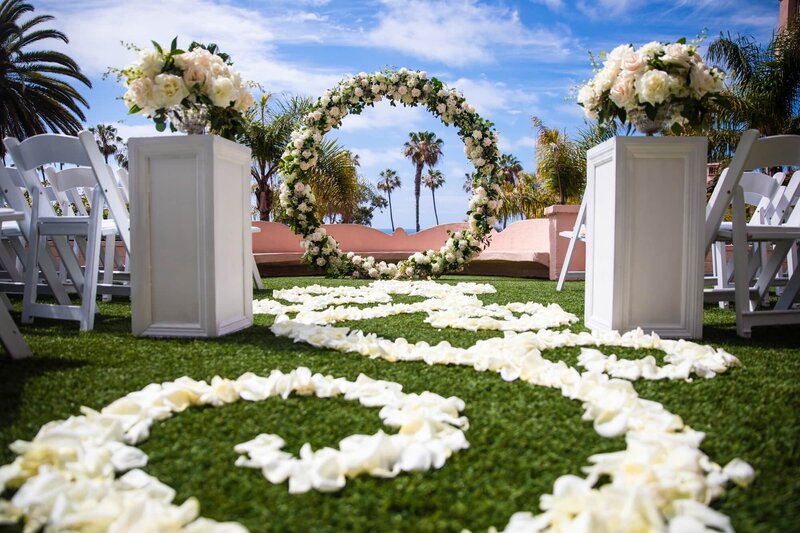 Wedding ceremony setup with floral bouquets, a floral arch, palm trees, grass, and flower petals on the ground at La Valencia Hotel in San Diego.