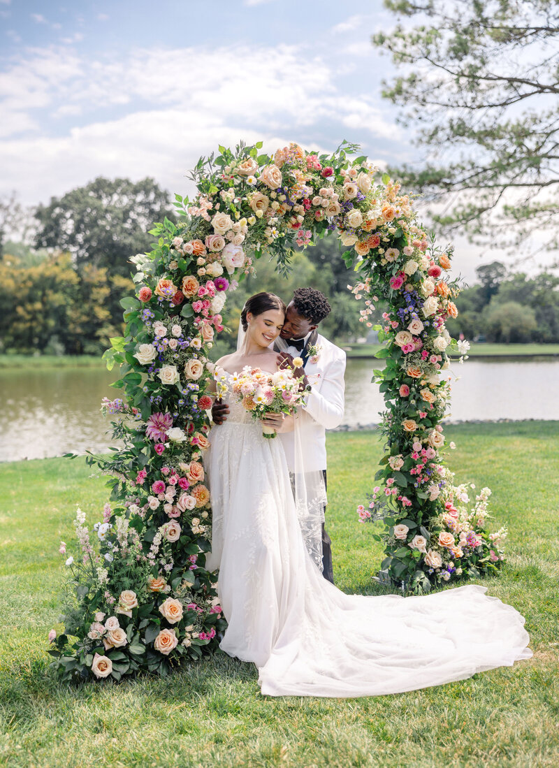 A bride and groom stand under a vibrant, flower-adorned arch, sharing a tender moment during their outdoor wedding. The scene is set by a tranquil lake and lush greenery under a partly cloudy sky. The bride holds a bouquet of colorful flowers.