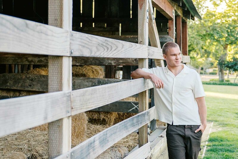 Teen boy wearing a white button up shirt and black jeans while leaning against a barn