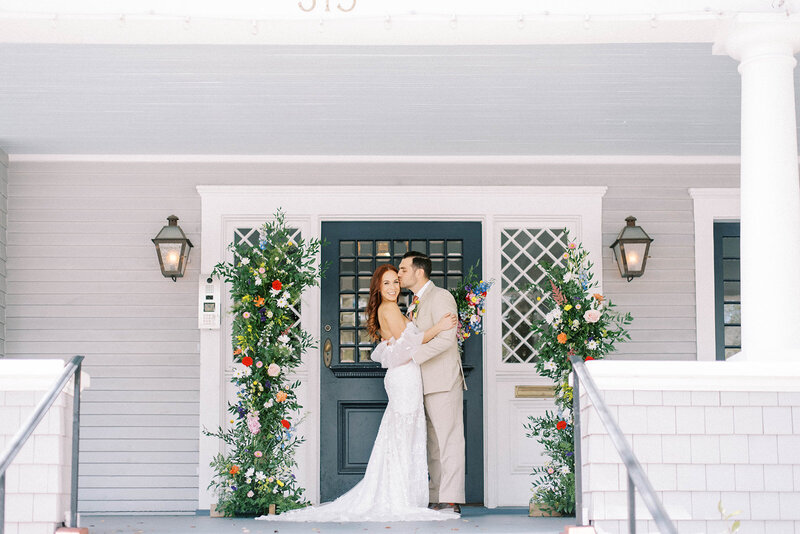 groom kissing his bride on the forehead as she turns to look at the camera while they stand in fron tog a dark blue door on a large porch on the Orlo wedding venue with bright florals decorating the space around the door