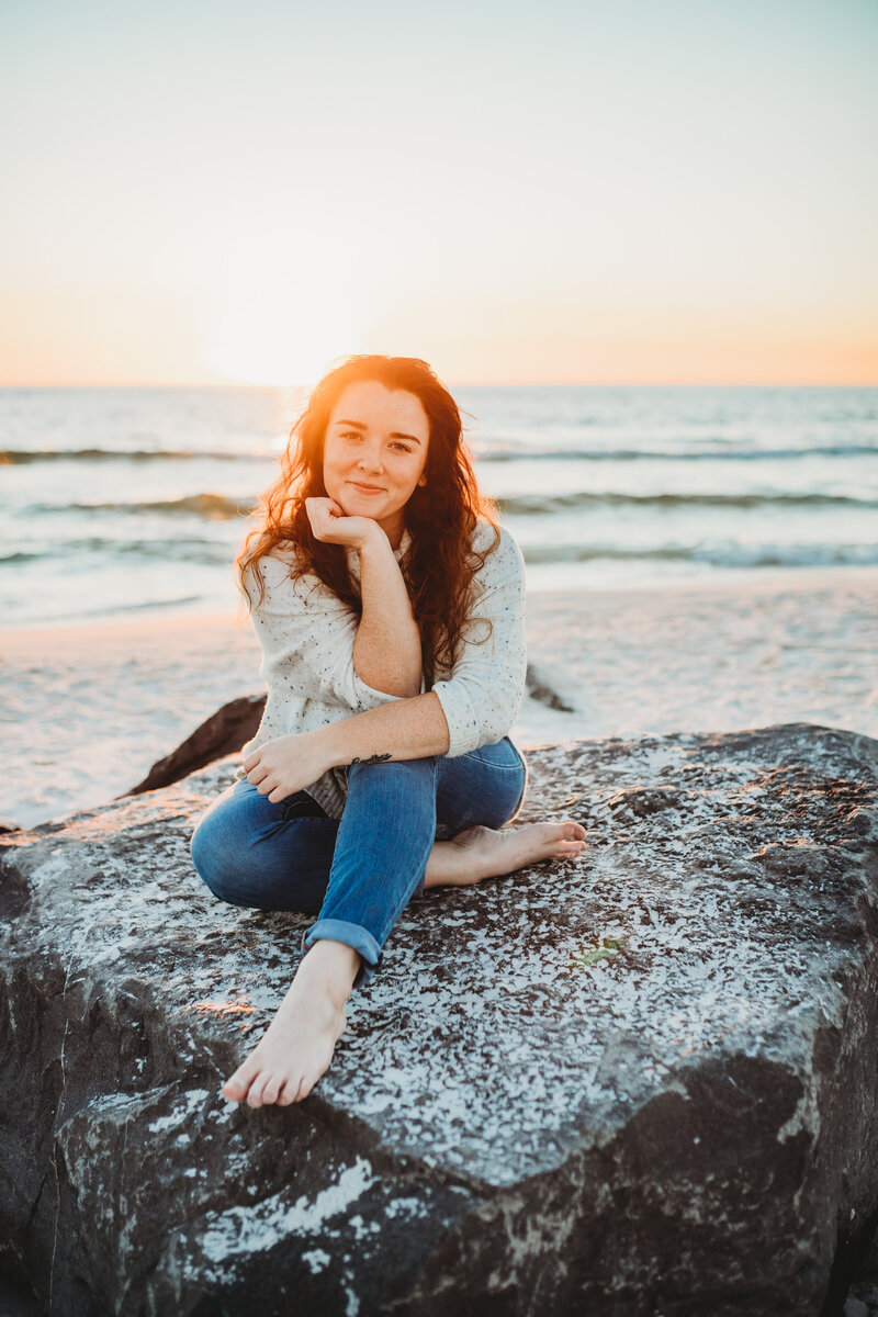 portrait of Brittney Stanley sitting on rock in Panama City Beach, smiling at camera wearing white sweater and rolled up jeans