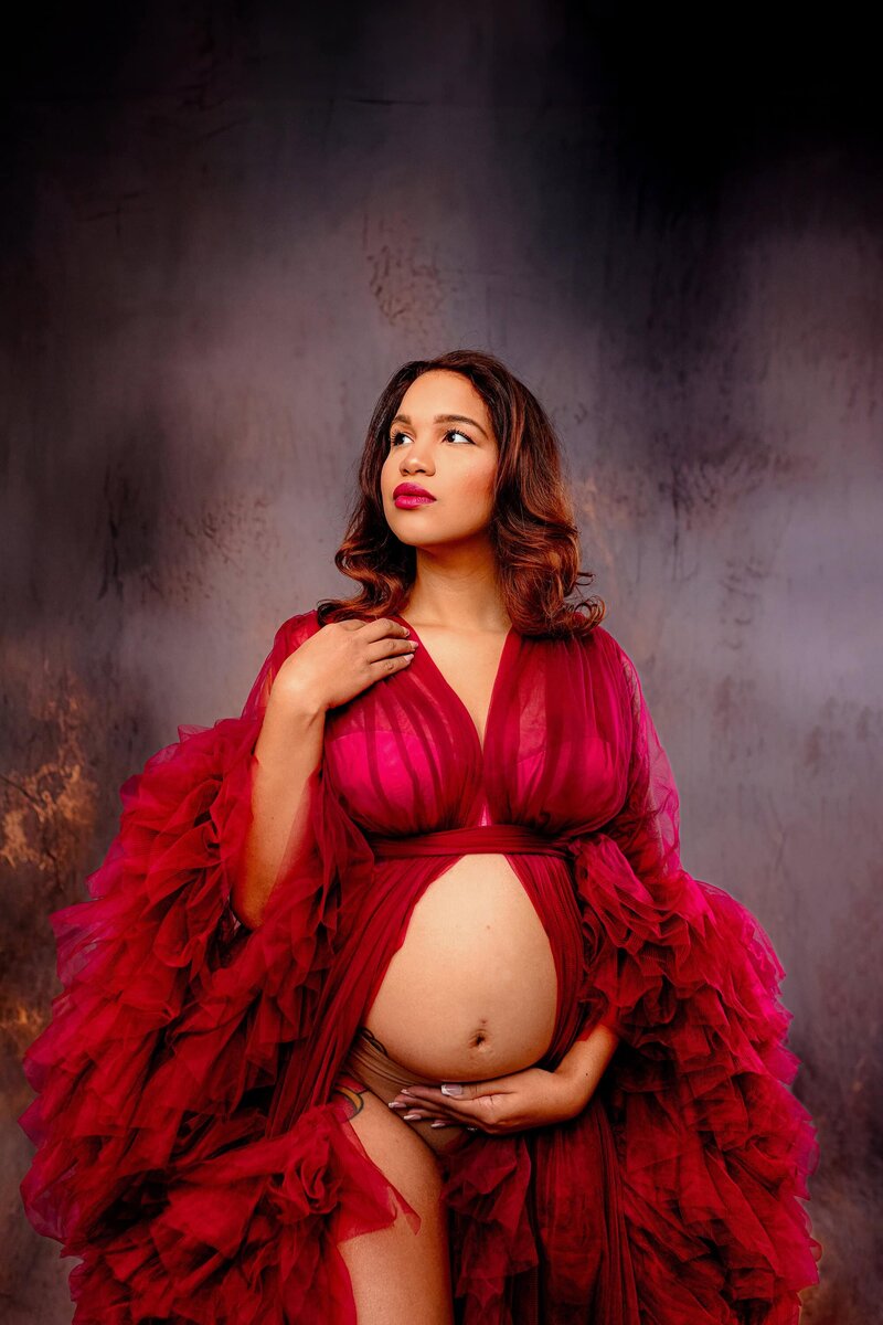 Pregnant mom touches her shoulder with one hand cradling her belly bump with the other looking off camera in a studio session