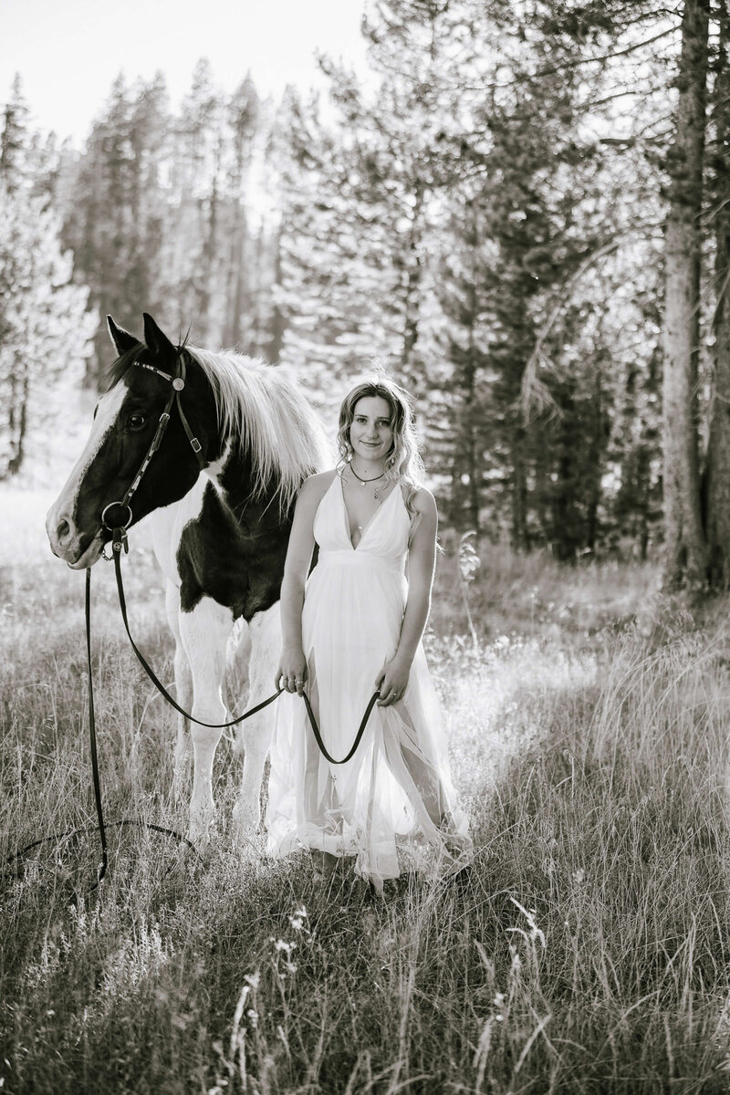 Senior girl portrait session in Truckee a black and white image of hre standing beside her horse in a white long dress looking at camera