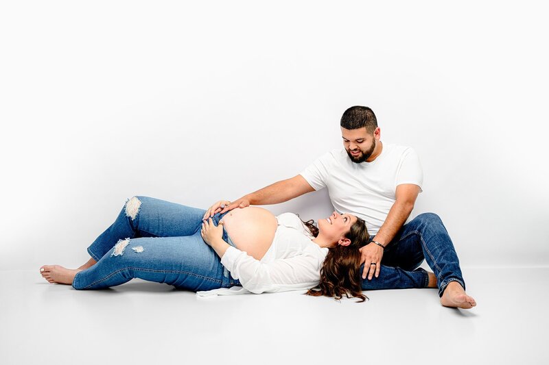 husband is dressed in jeans and a white shirt sitting with his expectant wife laying beside him with his hand on her belly