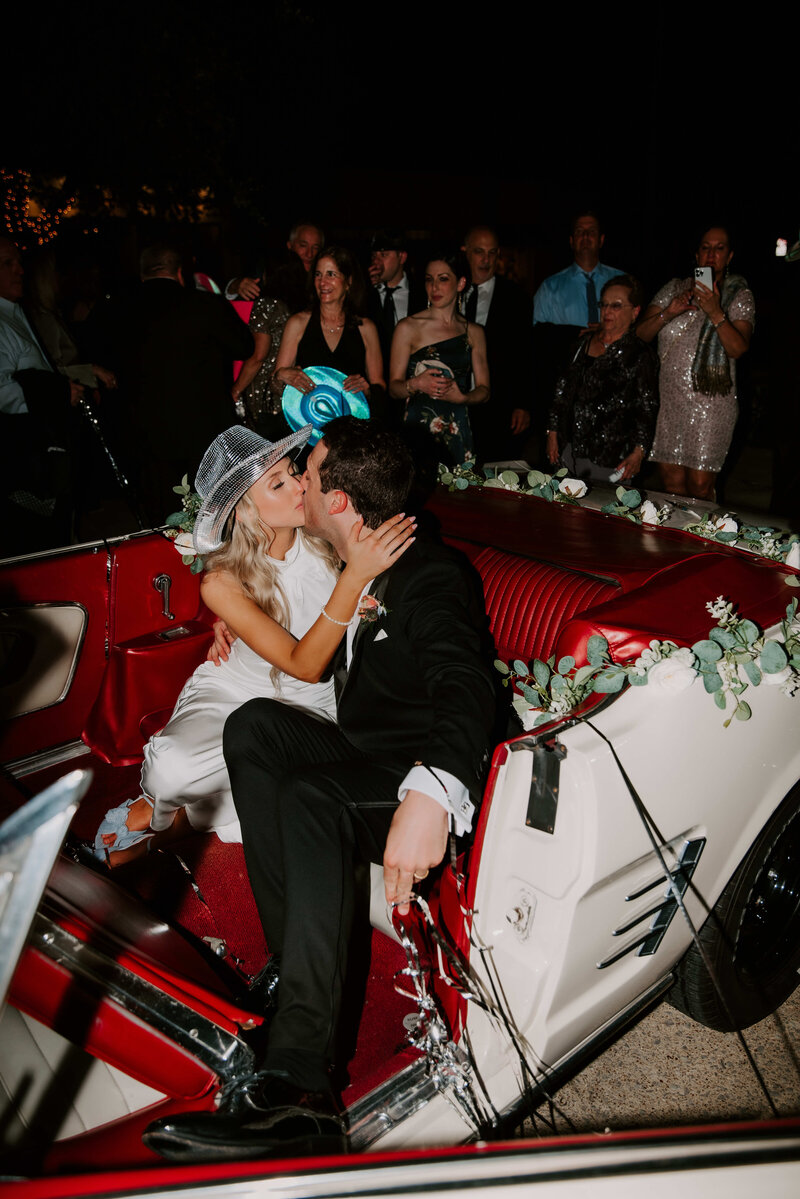 A couple is kissing in the backseat of a vintage white convertible with a red interior. The car is decorated with white flowers and green foliage along the back. The woman, wearing a silvery cowboy hat, is on the left, facing and kissing the man, who is wearing a black suit on the right. In the background, several people are visible, watching the couple.