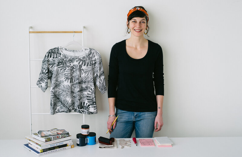 Image shows textile designer and artist Skye McNeill with a set of materials and samples for block printing. Skye wears a black shirt, jeans, and an orange headscarf and stands next to a block printed tee shirt she printed and sewed. On the table are block printing inks, rollers, blocks, and tools.