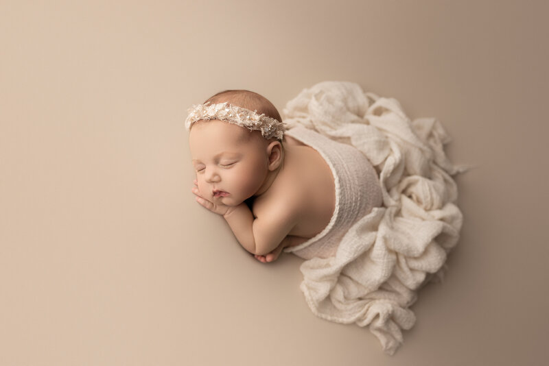Main Line Philadelphia newborn baby photoshoot: Baby girl lies atop of an ecru-coloured fabric and draped in cream-coloured fabric that is ruffled behind her. She is wearing a cream-coloured headband. Baby is lying on her belly with one hand under her cheek. Captured by best newborn photographer in Philadelphia Main Line Katie Marshall.