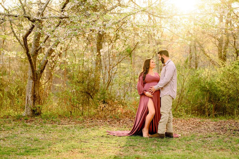 Expectant couple is posed together with hands on the new mom's belly and flowers from the spring trees behind them