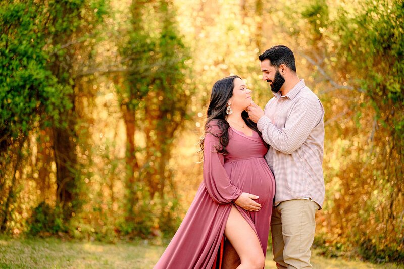 A father to be places his hand gently on his wife's chin as they look towards each other she has one hand on her belly and they're smiling together in an outdoor session