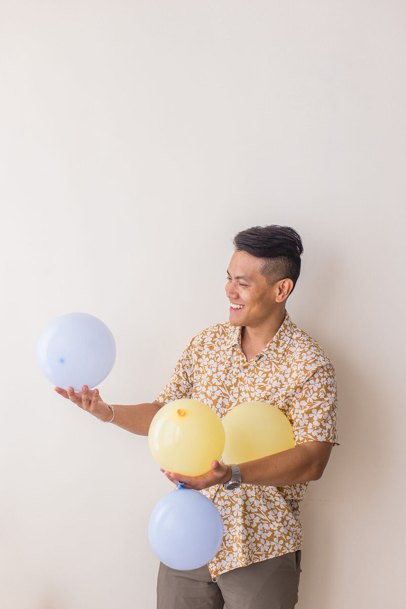 Sho Dewan, young Asian man, career coach and founder of Workhap is standing against a white wall, holding air balloons in his hands and laughing