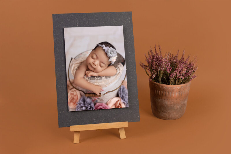A framed photo by a Jacksonville newborn photographer features a sleeping baby in a flower headband, displayed on a small wooden easel beside a potted purple heather, set against a warm brown background.