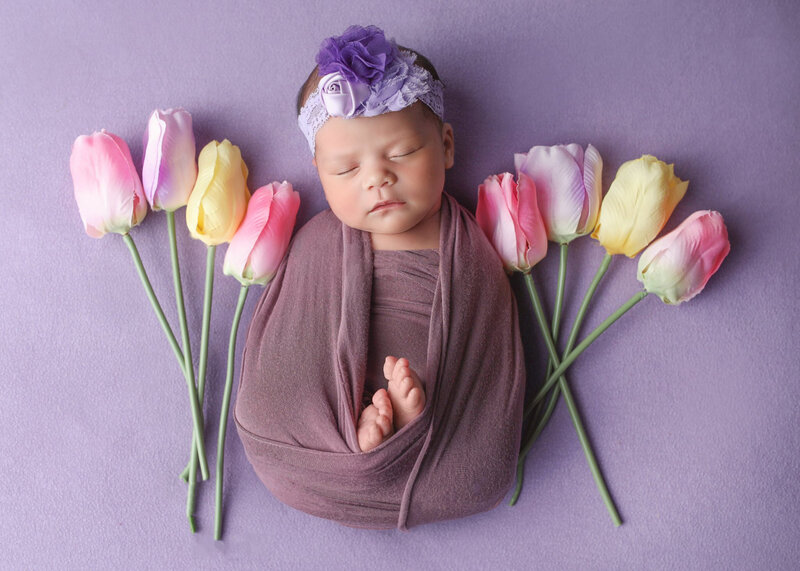 Brand new infant baby sleeping peacefully on purple beanbag blanket surrounded by pretty tulips with purple floral headband on.