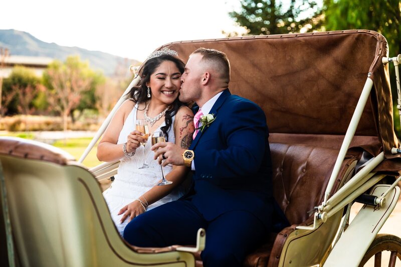 Bride and groom sharing a kiss in a horse-drawn carriage with champagne after their wedding ceremony at Galway Downs in Temecula