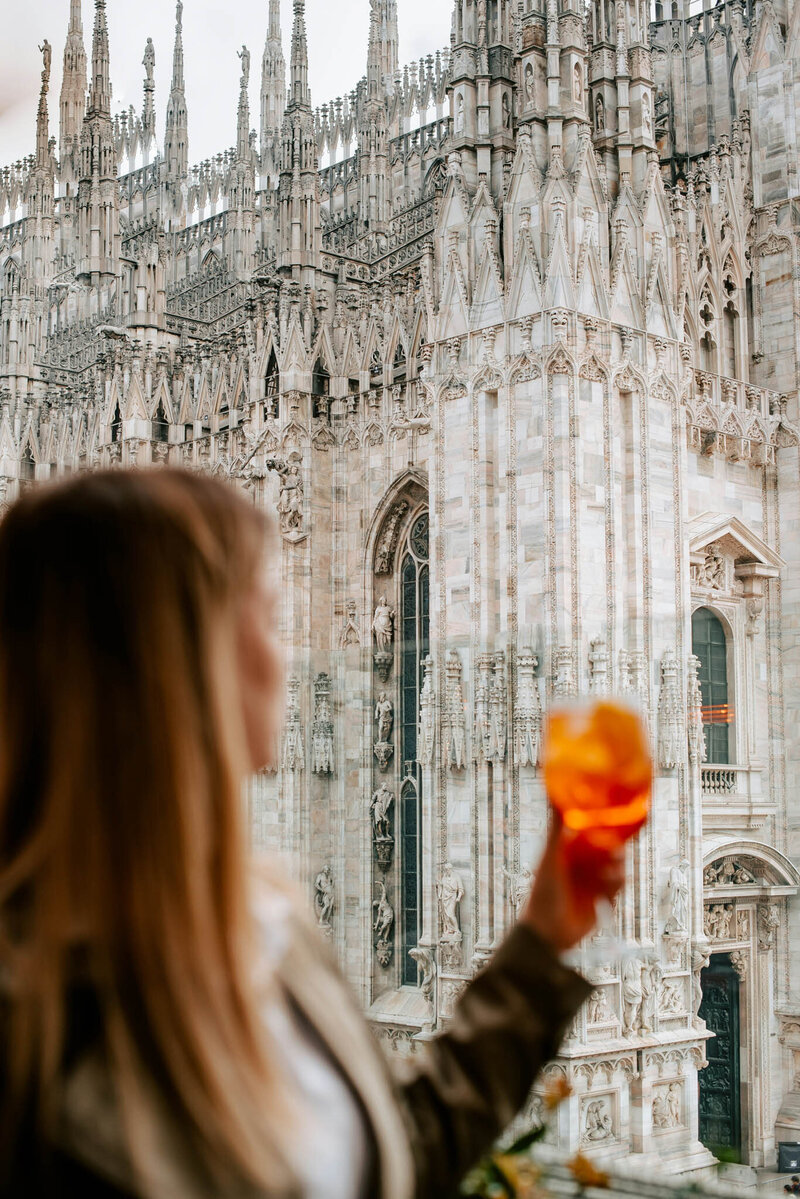 A person holds an orange drink while looking at the ornate facade of a Gothic cathedral with intricate spires and sculptures. The focus is on the cathedral, while the person in the foreground is blurred.
