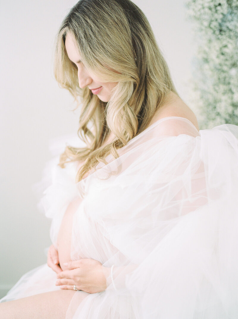 A blonde mother in a tulle dress, sitting down and holding her pregnant belly, photographed by Edmonton Newborn Photographer, Kahla Kristen Photography