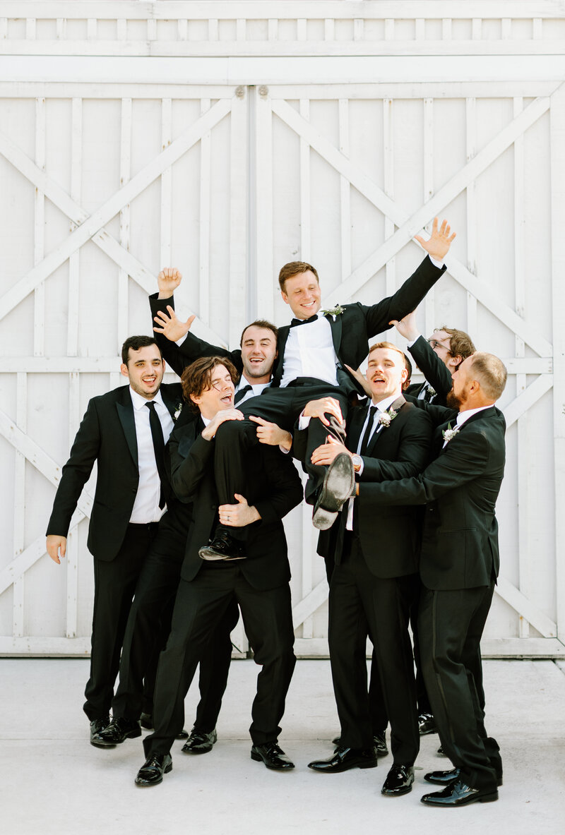 Groomsmen wearing classic black and white tuxes lifting groom in the air outside of barn doors at White Dove Barn