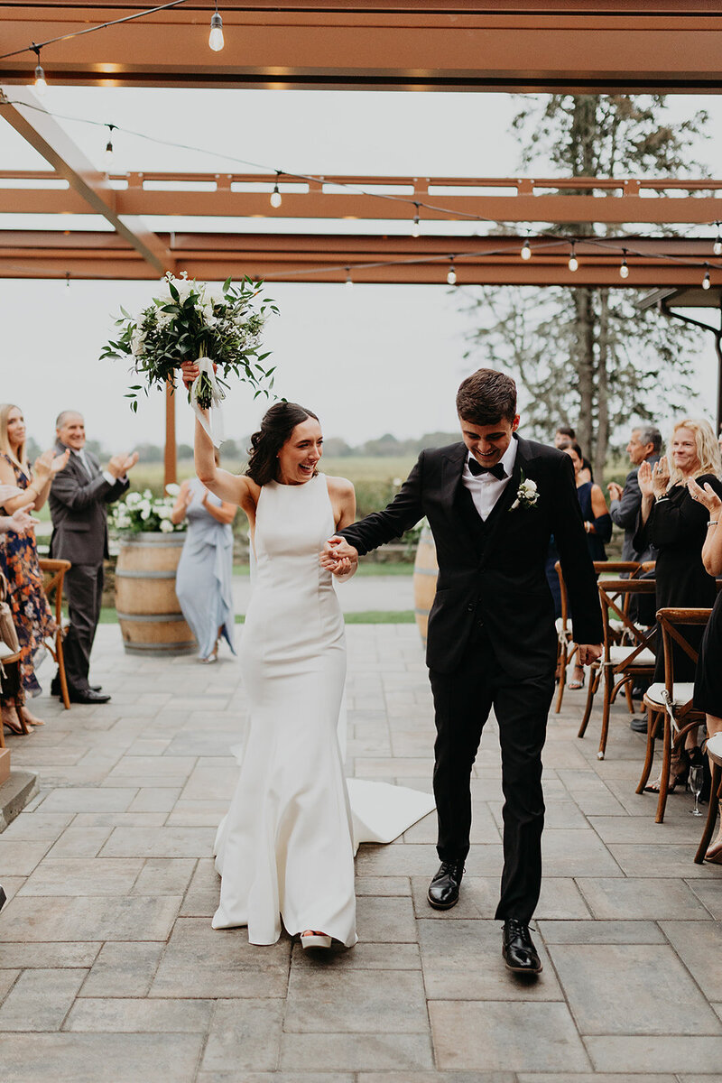 Capturing the joyous wedding moment at the vineyard, the bride and groom walk hand in hand from the altar, radiating happiness. The bride adds a playful touch, waving her bouquet with delight.