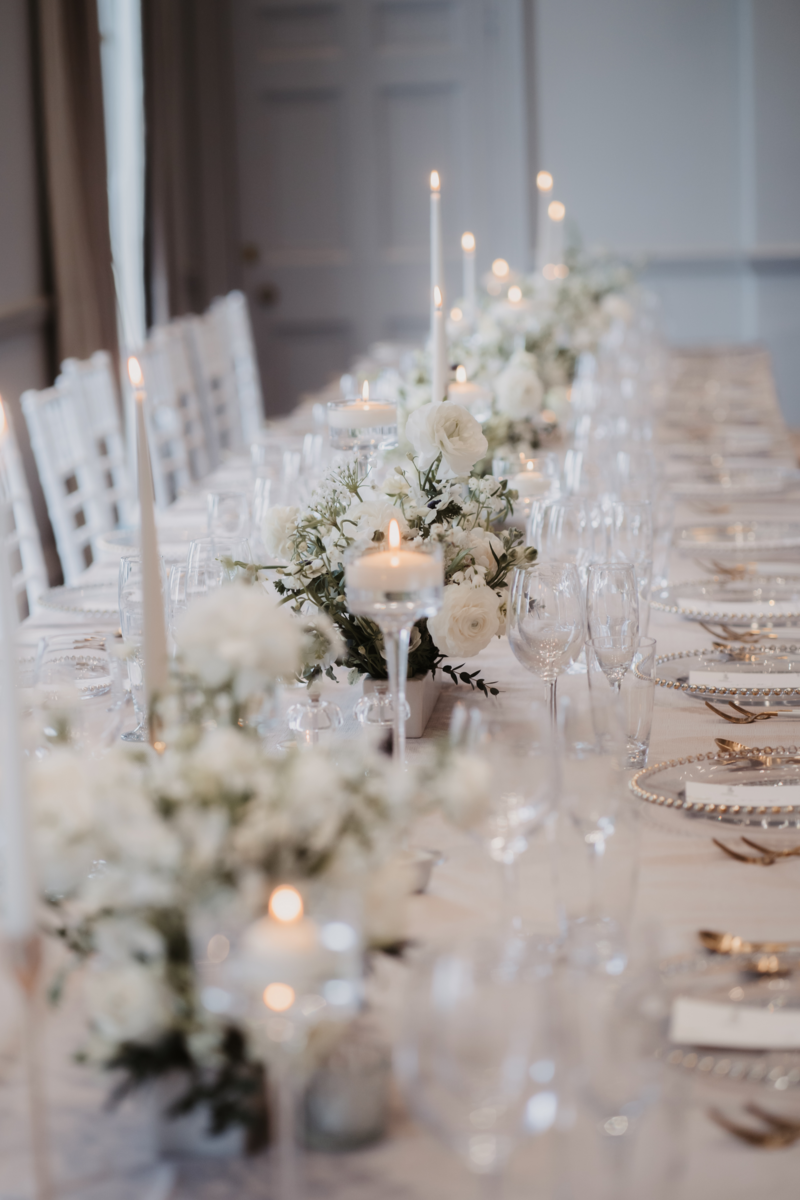 Wedding venue reception table sophisticated  white  flowers  in runner meadows with glassware and candles , charger plates, neutral table linen